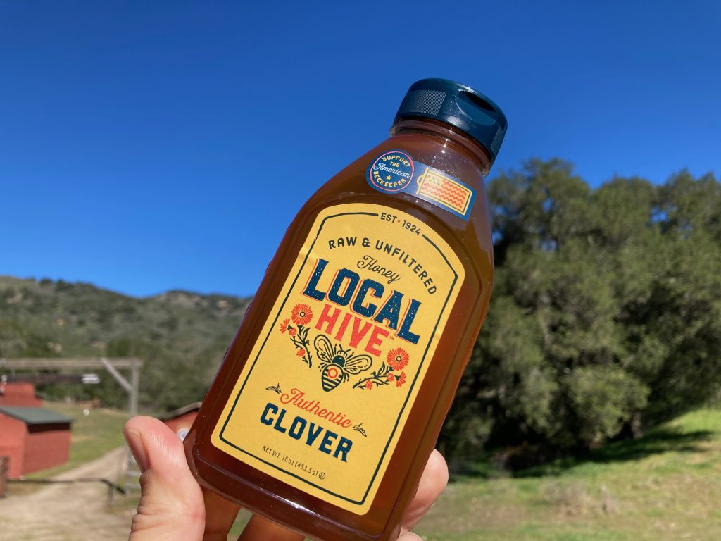 Hand holding local live honey brand of a jar of their raw unfiltered clover honey with mountains and blue sky in the background
